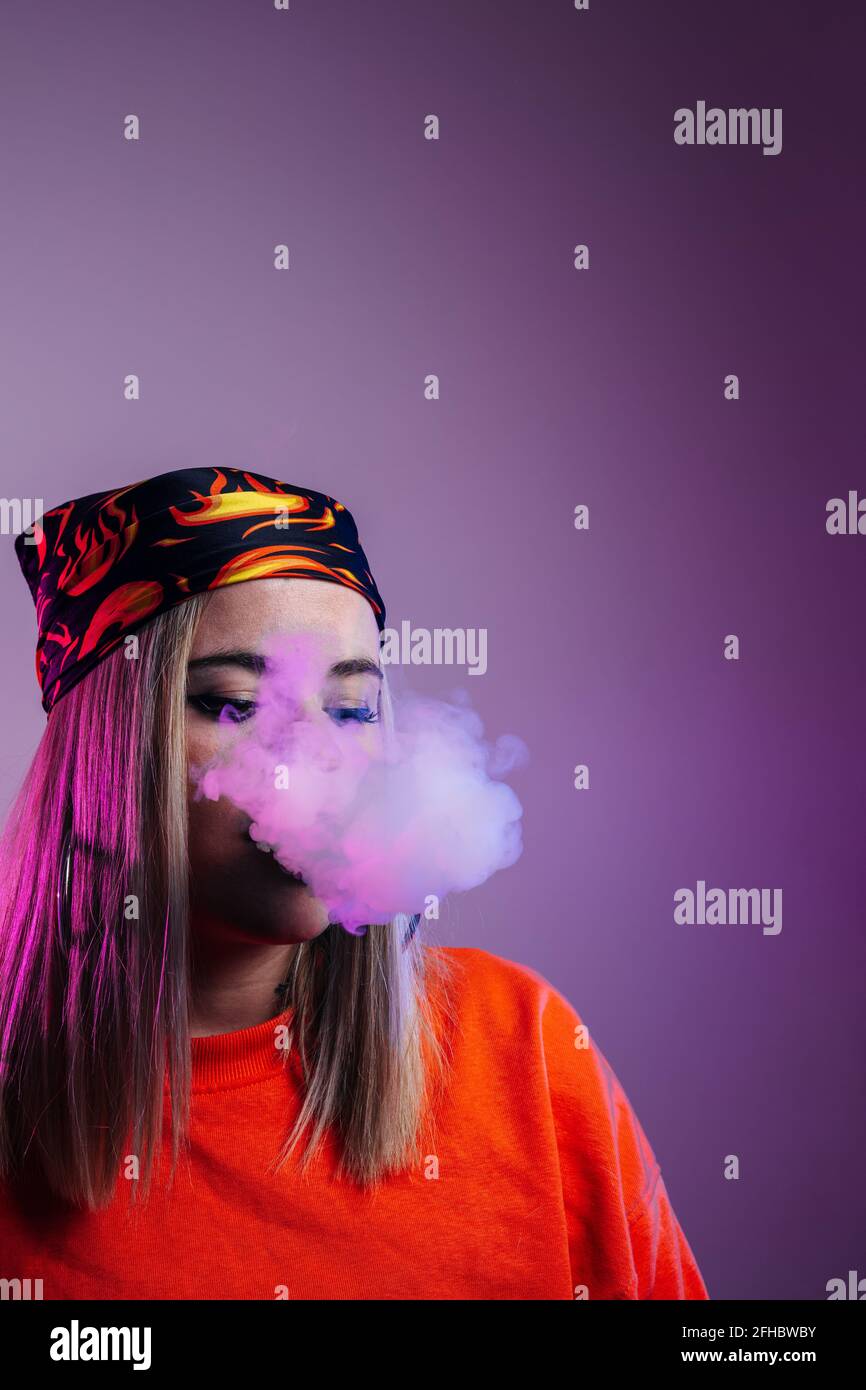 Side view of cool female in street style outfit smoking e cigarette and exhaling smoke through nose on purple background in studio with pink neon illu Stock Photo