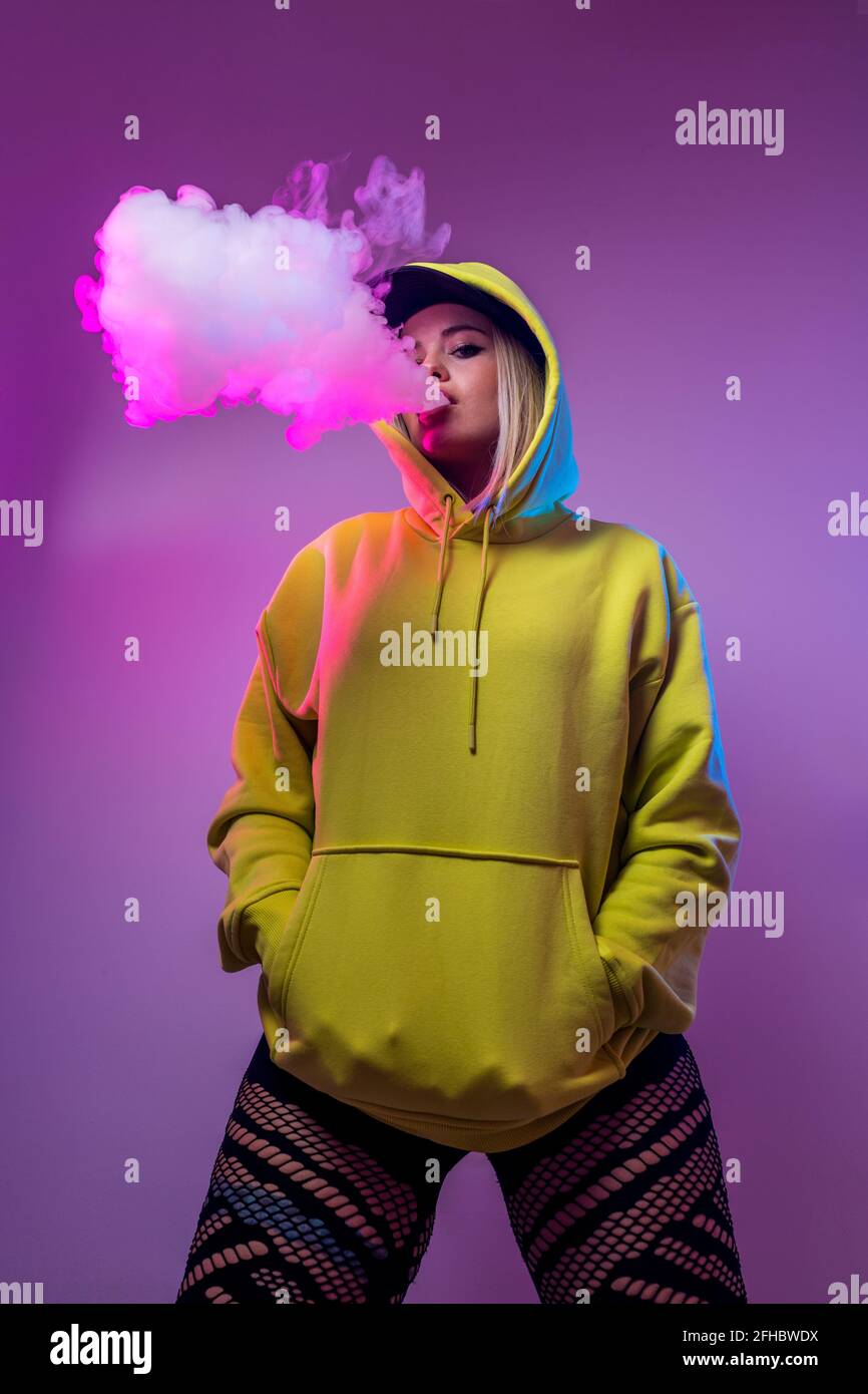 From below confident female hipster in hoodie smoking e cigarette in studio on pink background looking at camera Stock Photo
