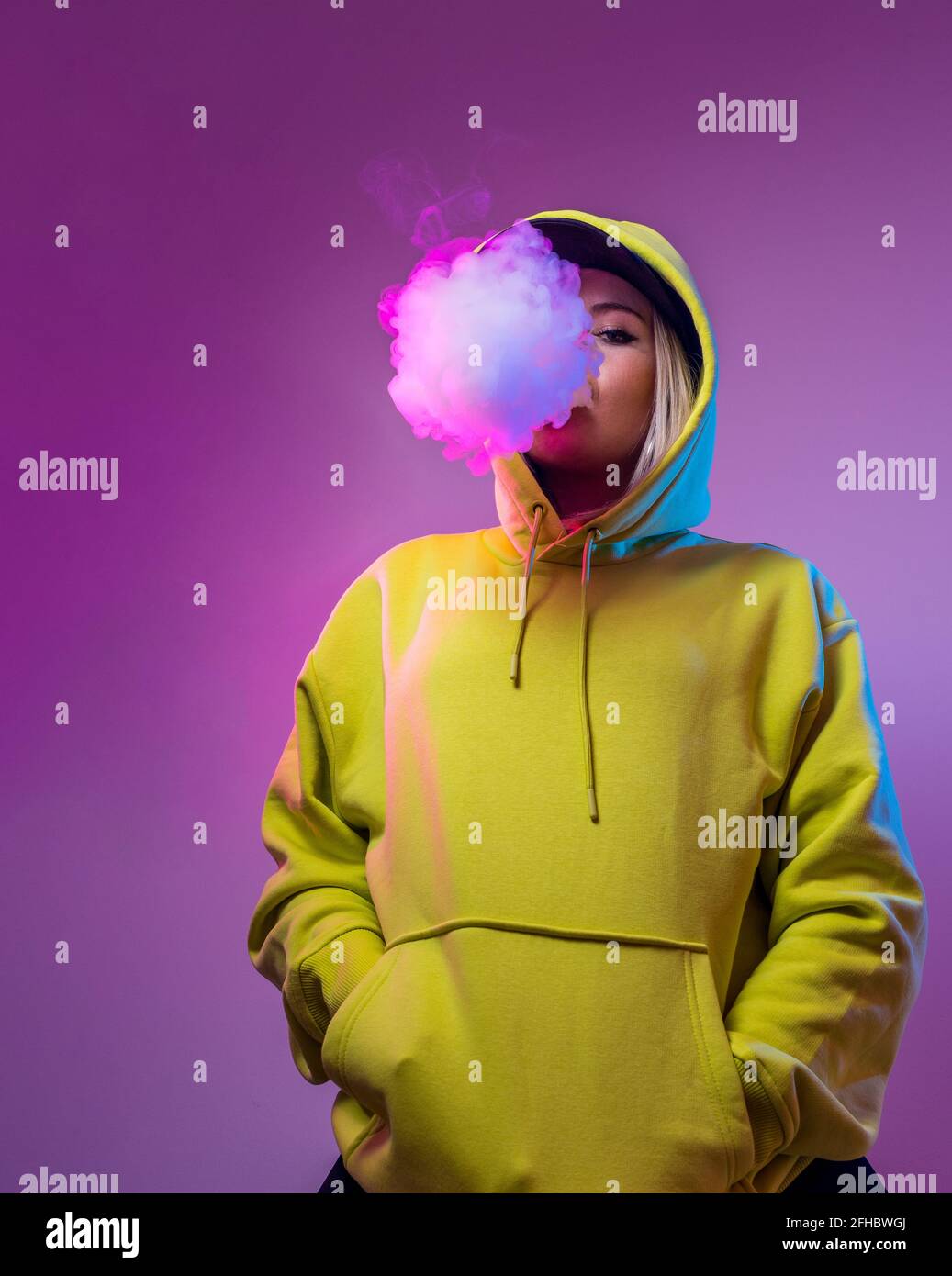 Confident female hipster in hoodie smoking e cigarette in studio on pink background and looking at camera Stock Photo