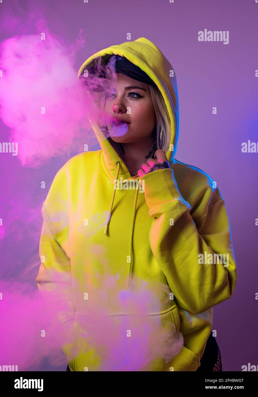 Confident female hipster in hoodie smoking e cigarette in studio on pink background and looking away Stock Photo