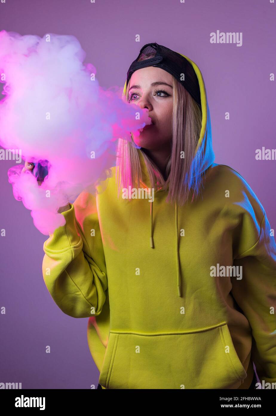 Confident female hipster in hoodie smoking e cigarette in studio on pink background and looking away Stock Photo
