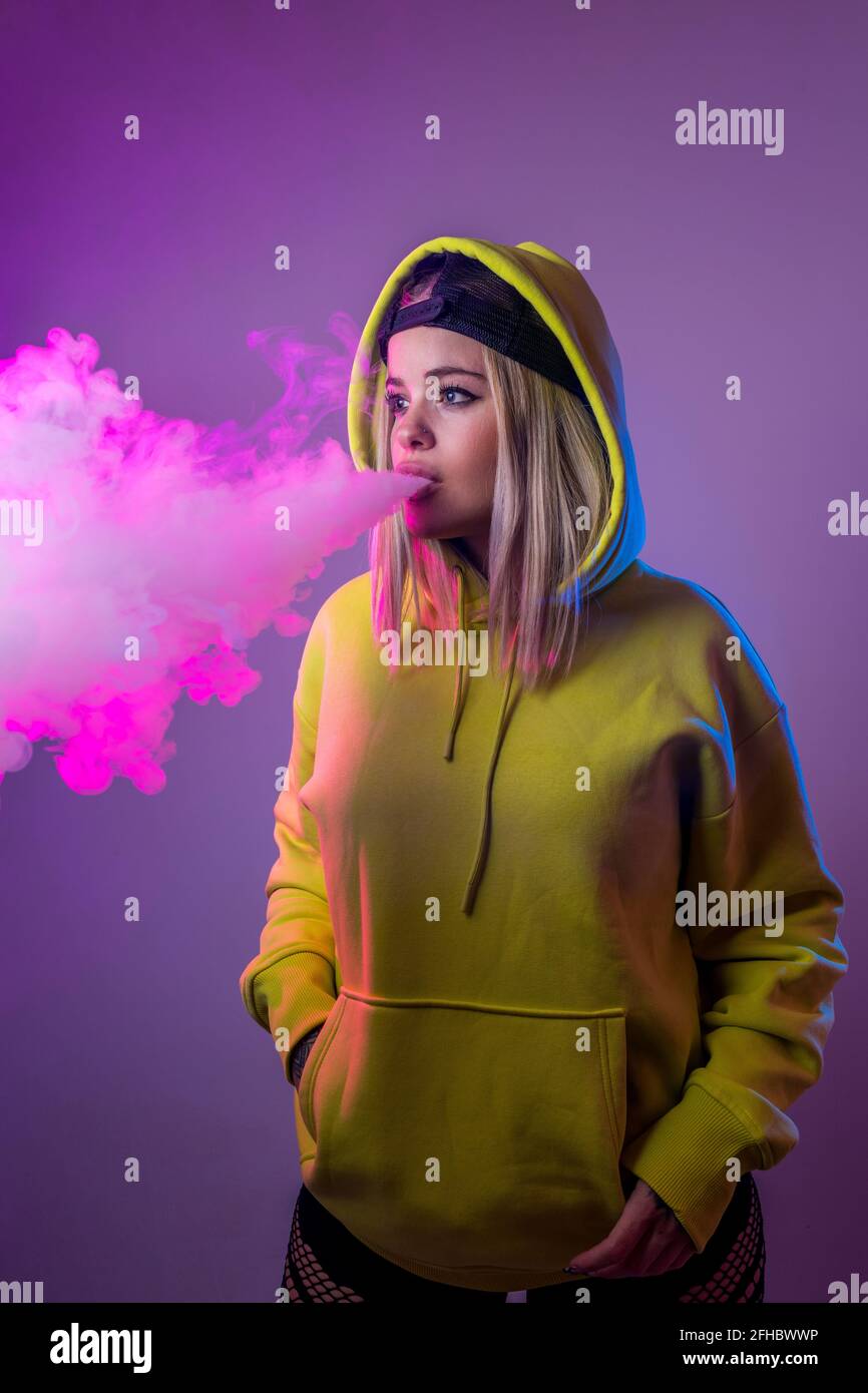 Confident female hipster in hoodie smoking e cigarette in studio on pink background and looking away Stock Photo