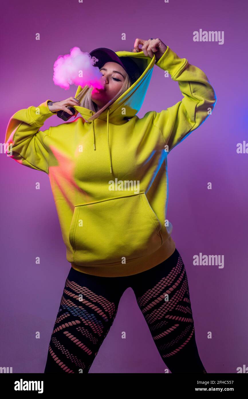 From below confident female hipster in hoodie smoking e cigarette in studio on pink background Stock Photo