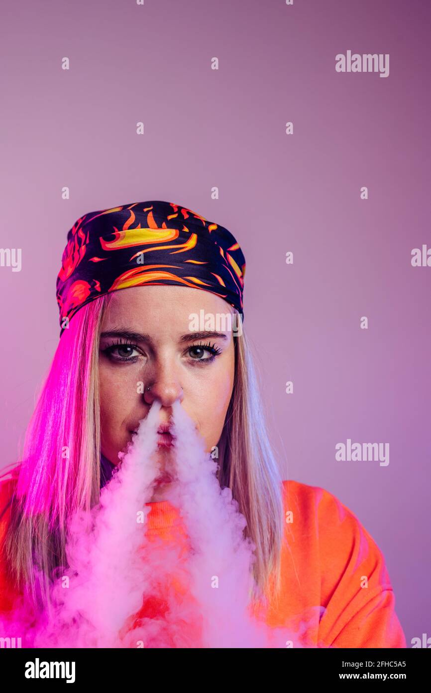 Cool female in street style outfit smoking e cigarette and exhaling smoke through nose on purple background in studio with pink neon illumination Stock Photo