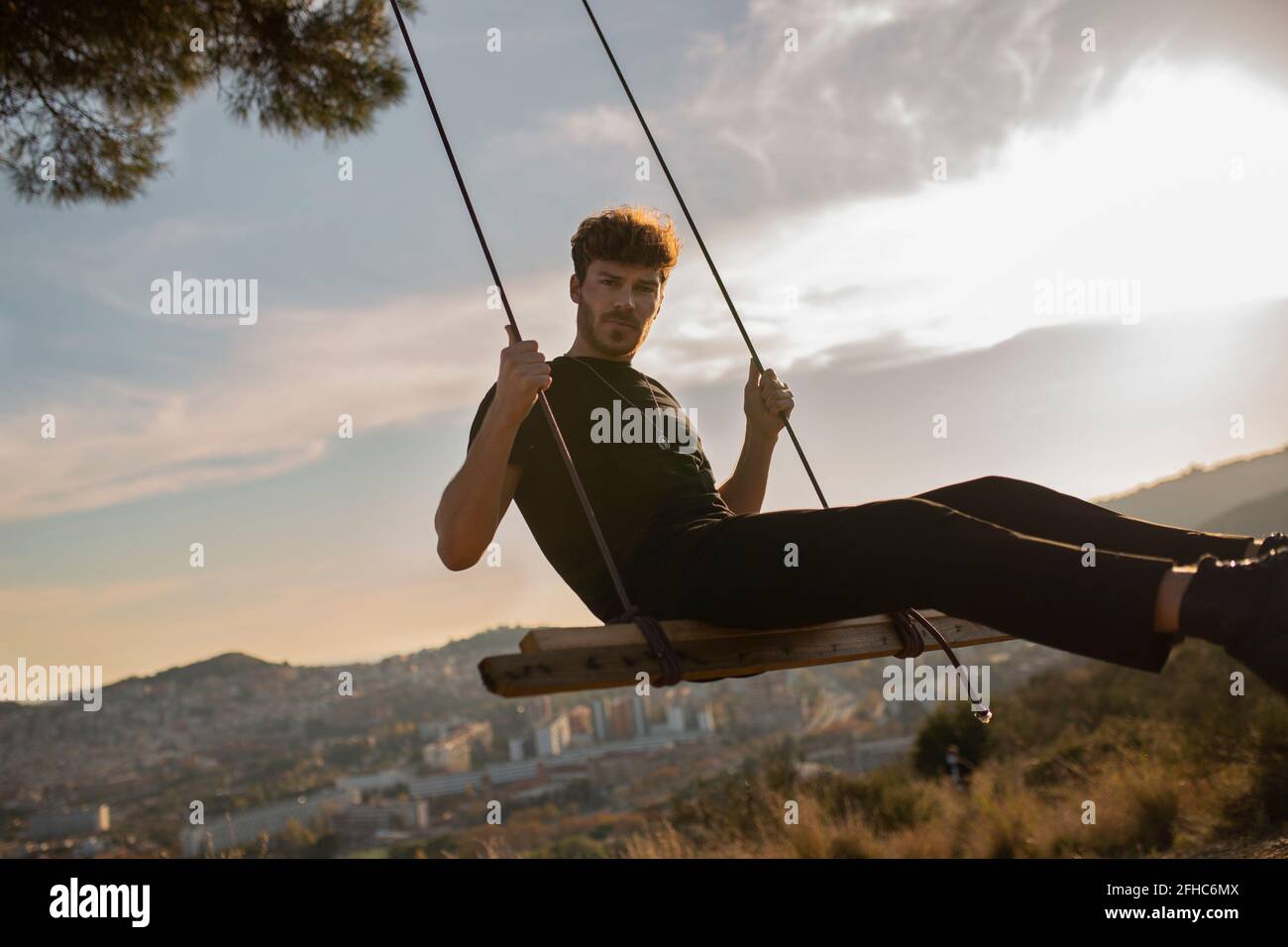 Bearded male in black apparel looking at camera while sitting on swing against mountains in town at sundown Stock Photo
