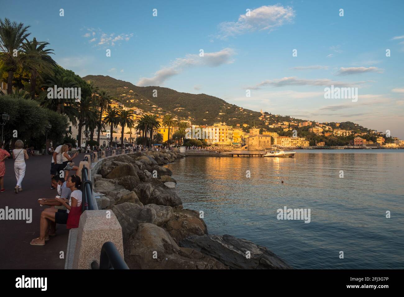 People are enjoying the 'lungomare' of Rapallo during summer. Rocks border the bay, while palm trees border the promenade. Stock Photo