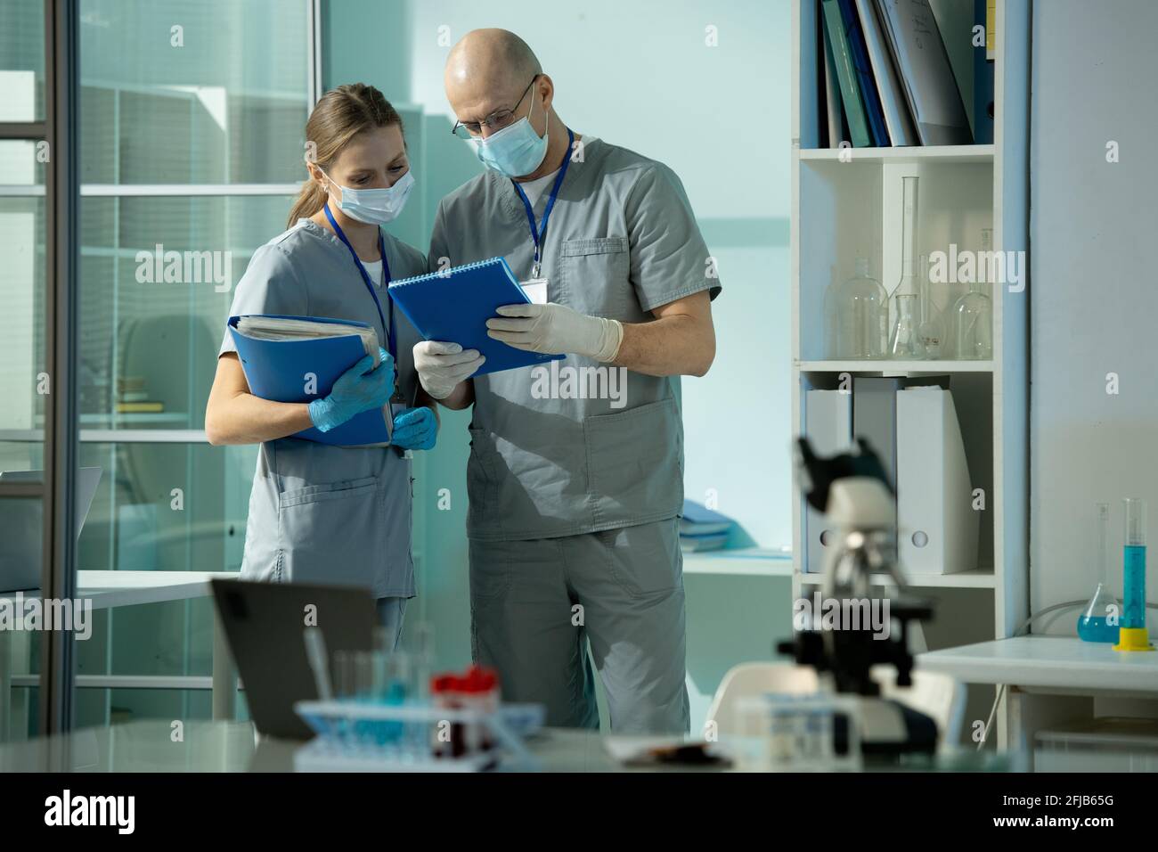 Busy medical colleagues in scrubs and surgical mask standing in laboratory room and reading records of patient Stock Photo