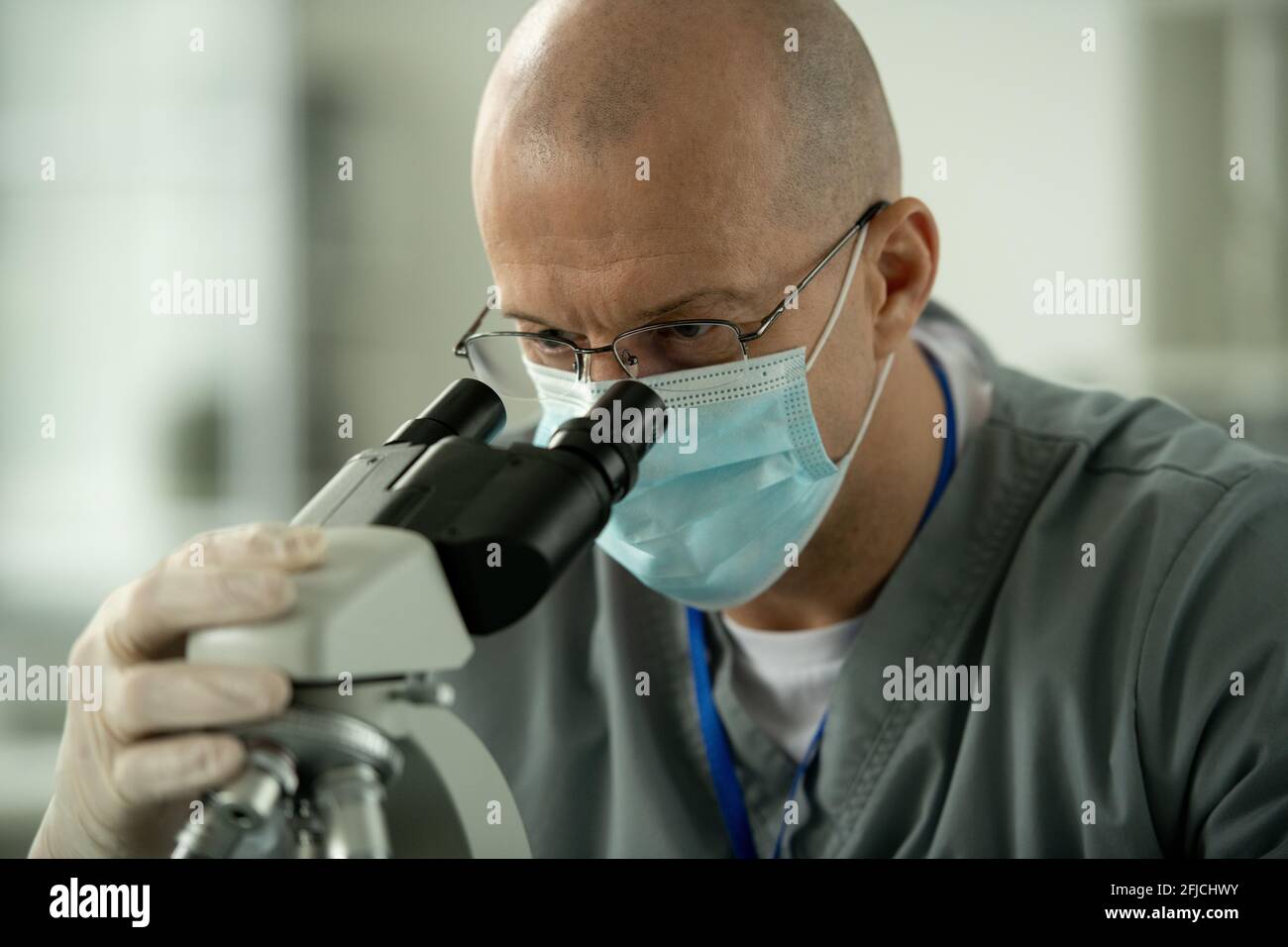Concentrated lab worker in glasses and mask using microscope while analyzing sample in laboratory Stock Photo