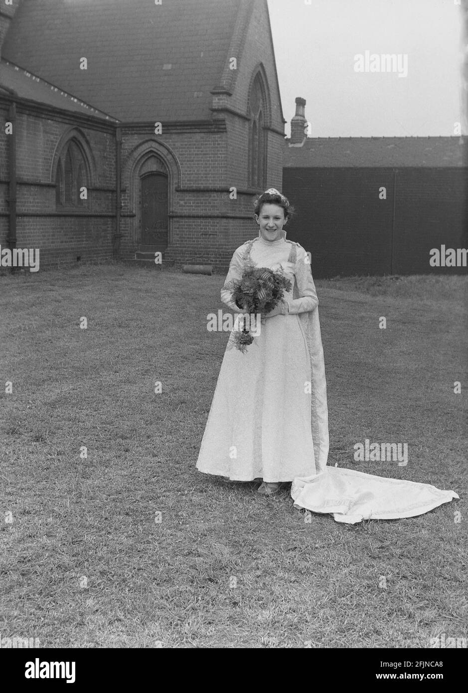 1956, historical, outside in the grounds of church, a young girl in her gown and holding a flower bouquet stnading for a photograph before the May Day carnival and the crowing of the town's May Queen. An ancient festival celebrating the arrival of Spring, May Day involved the crowning of a May Queen and dancing around a Maypole, activities that have taken place in England for centuries. Selected from the girls of the area, The May Queen would start the procession of floats and dancing. In the industrialised North of England, the Church Sunday Schools often led the organisation. Stock Photo
