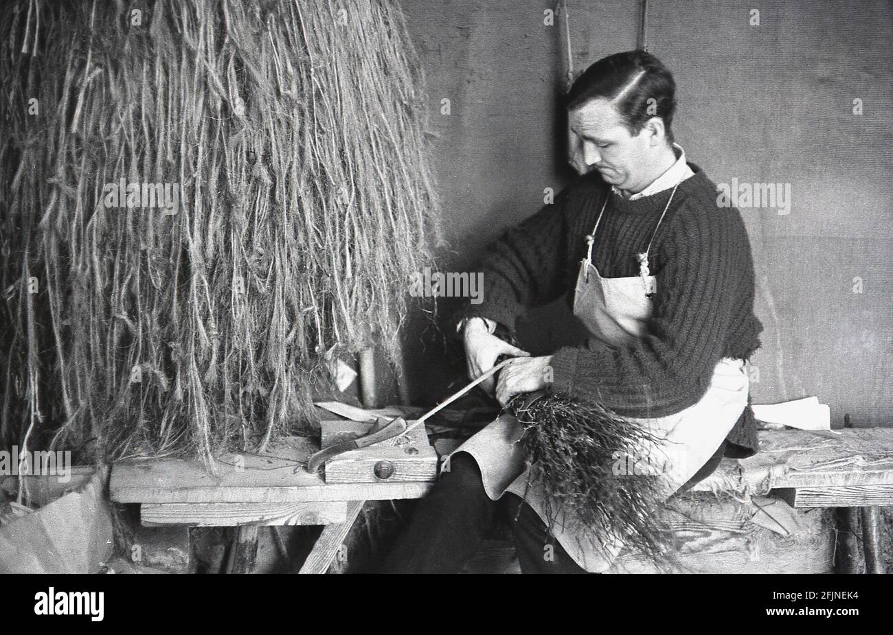 1950s, historical, a male artisan in his workshop making a broom, England, UK. An ancient craft, the making of brooms, also known as 'besoms' or 'besom making', consists of a bundle of tree twigs tied securely around a stick. Traditionally, besom brooms were made from twigs of the birch tree and a village broom squire and coppiceworker both produced them. Today, lIke many ancient rural crafts, this highly specialised artisan trade has almost completely died out and there are now fewer than five besom makers in the UK who make brooms on a commmerical basis. Stock Photo
