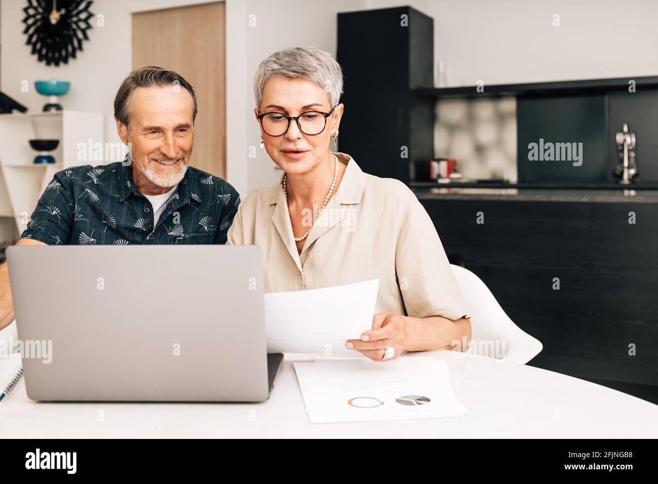 Senior retired couple at a table in dining room using a laptop computer. Two mature people looking at paperwork and discussing their finances. Stock Photo