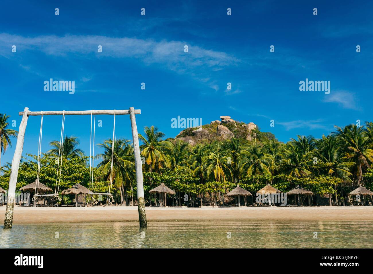 A tropical beach with a wooden swing in the water, a sandy beach on a paradise island, an empty beach with parasols near the coconut trees with a moun Stock Photo
