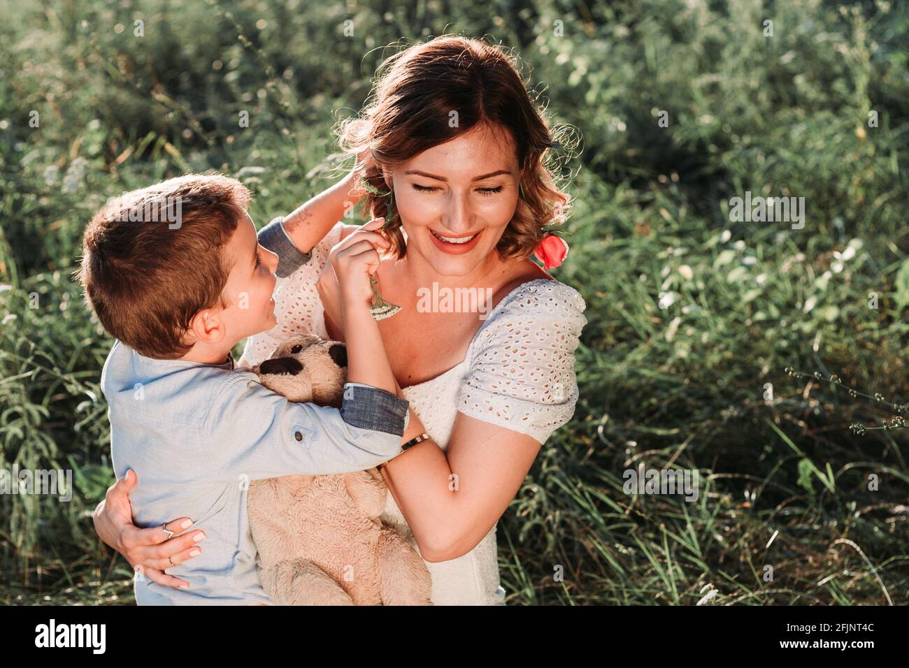 Beautiful young mom holding her blond son in her arms. He is putting flower in her hair. Outdoor photo. Mothers day Stock Photo
