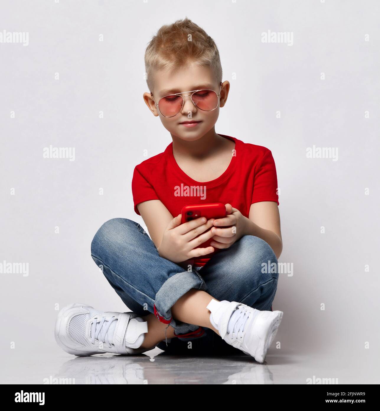 Stylish kid boy child in red t-shirt, jeans, sneakers and sunglasses sitting on floor with legs crossed looking at smartphone Stock Photo