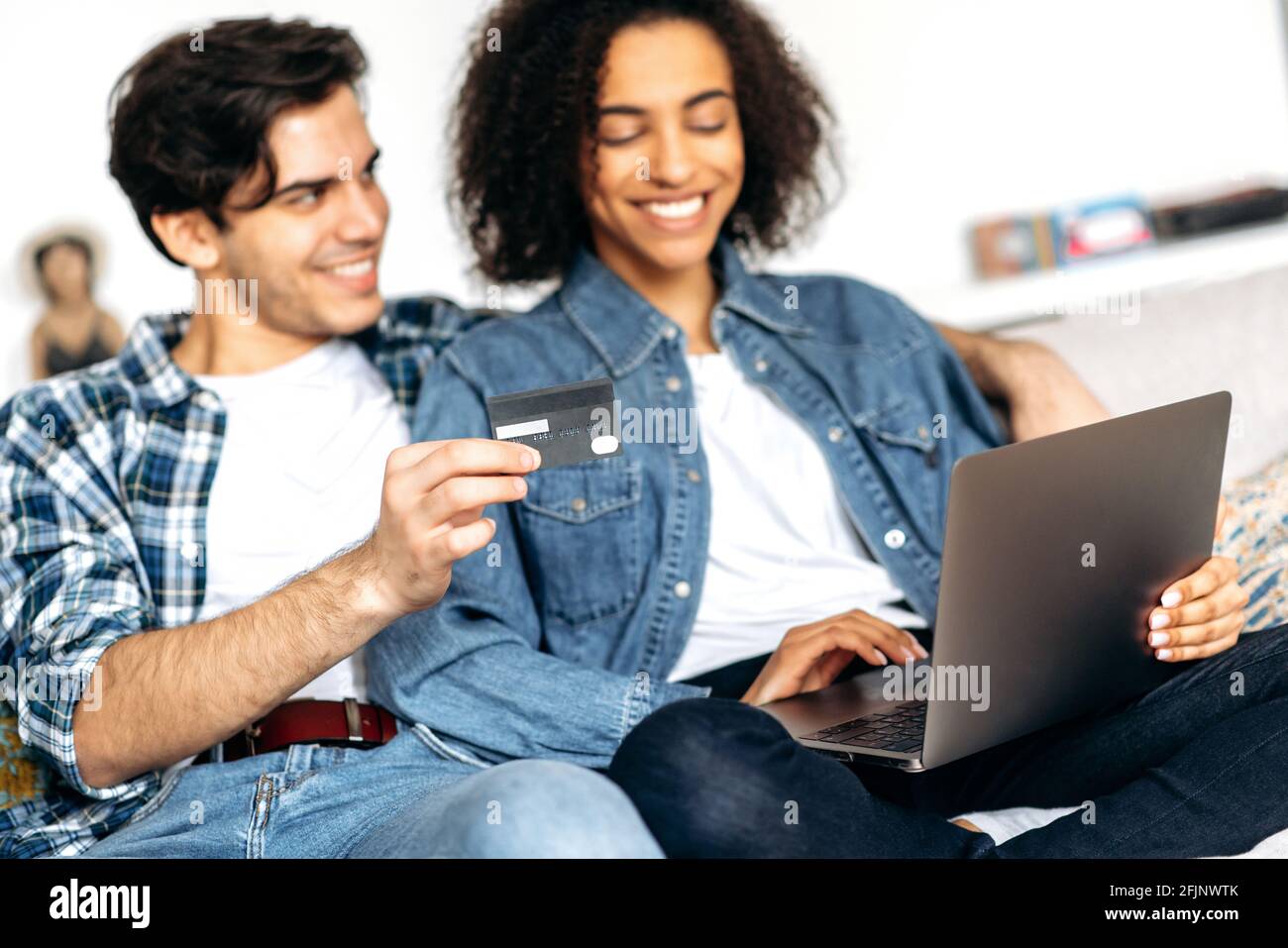Joyful young mixed race family couple, guy with girl sitting on sofa in living room, dressed in casual stylish clothes, shopping online using laptop and credit card, paying for goods, smiling Stock Photo