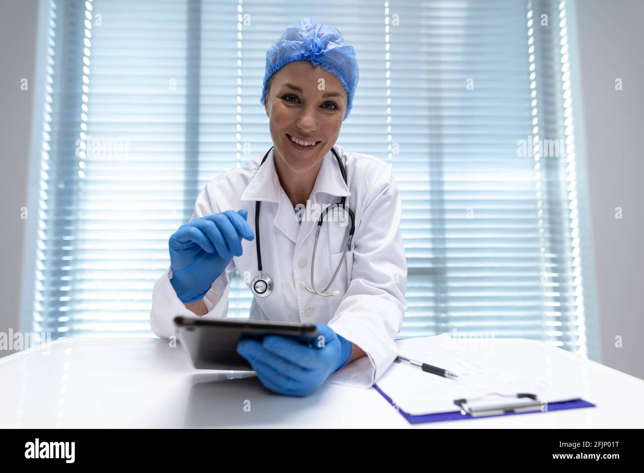 Caucasian female doctor at desk using tablet and smiling during video call consultation Stock Photo