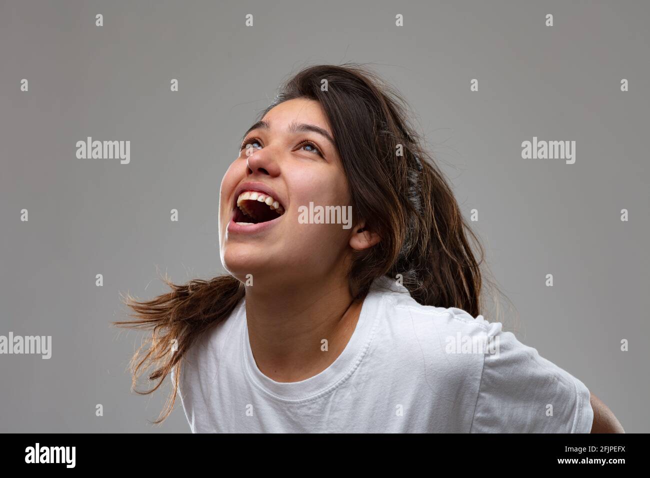 Exuberant young woman vocalising laughing and having fun or screaming in frustration with head tilted back and mouth open over grey Stock Photo