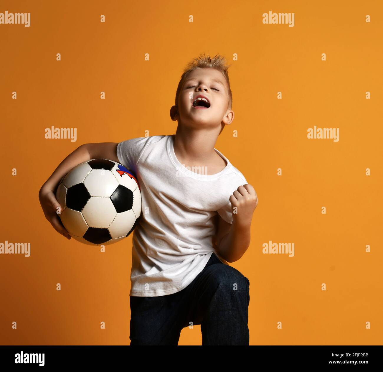 Happy schoolboy in white blank t-shirt and jeans stands holding soccer ball in hand and celebrates Goal Stock Photo