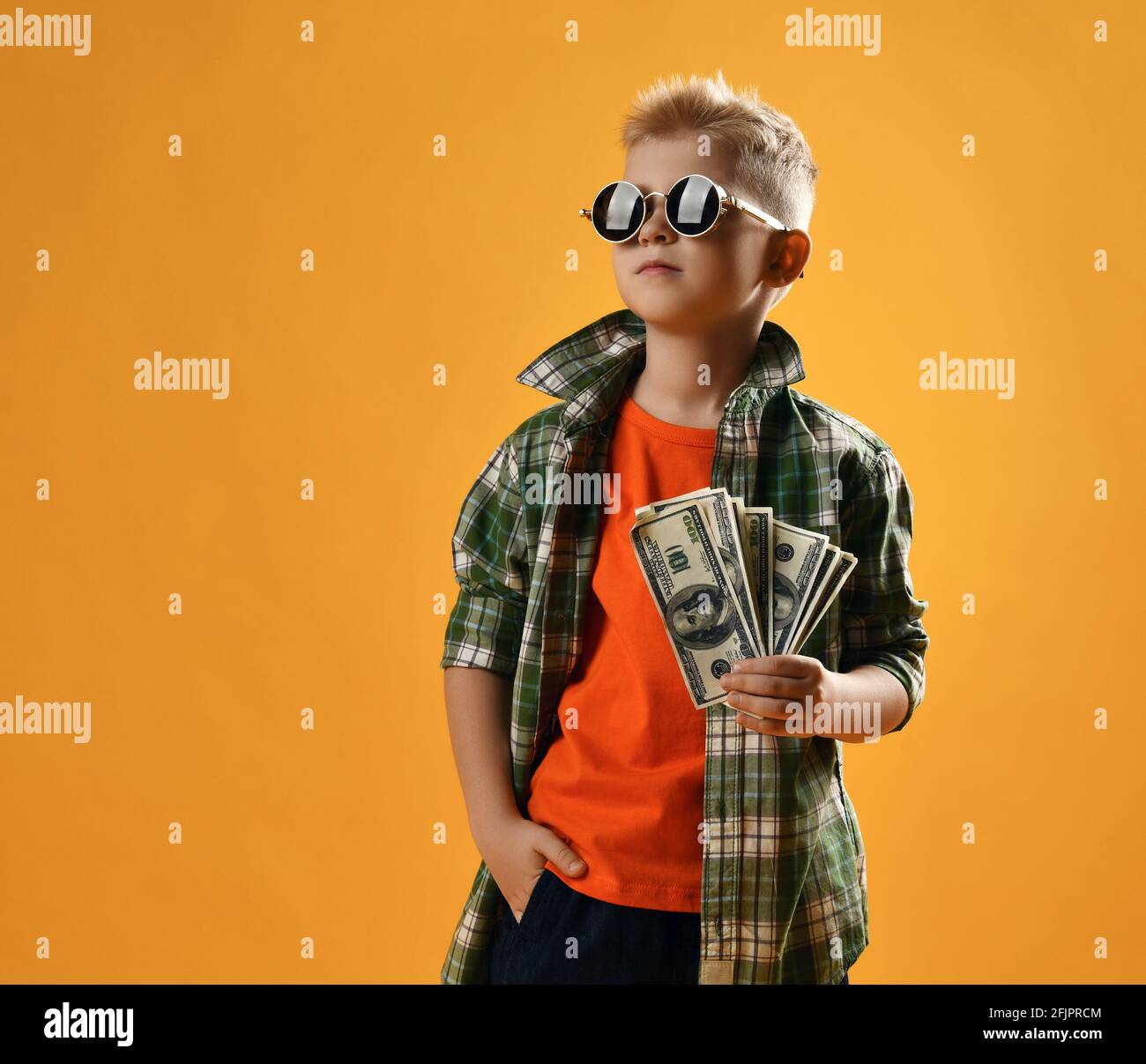 Calm self-confident rich teenager boy in round sunglasses, checkered plaid shirt and jeans stands holding fan of cash Stock Photo
