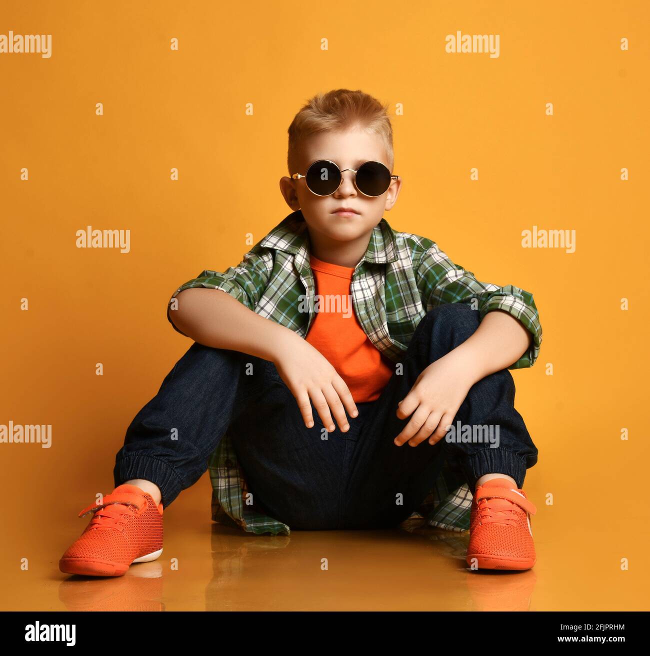 Young rock star, schoolboy teenager leader in round sunglasses, checkered shirt, orange t-shirt and jeans sits on floor Stock Photo