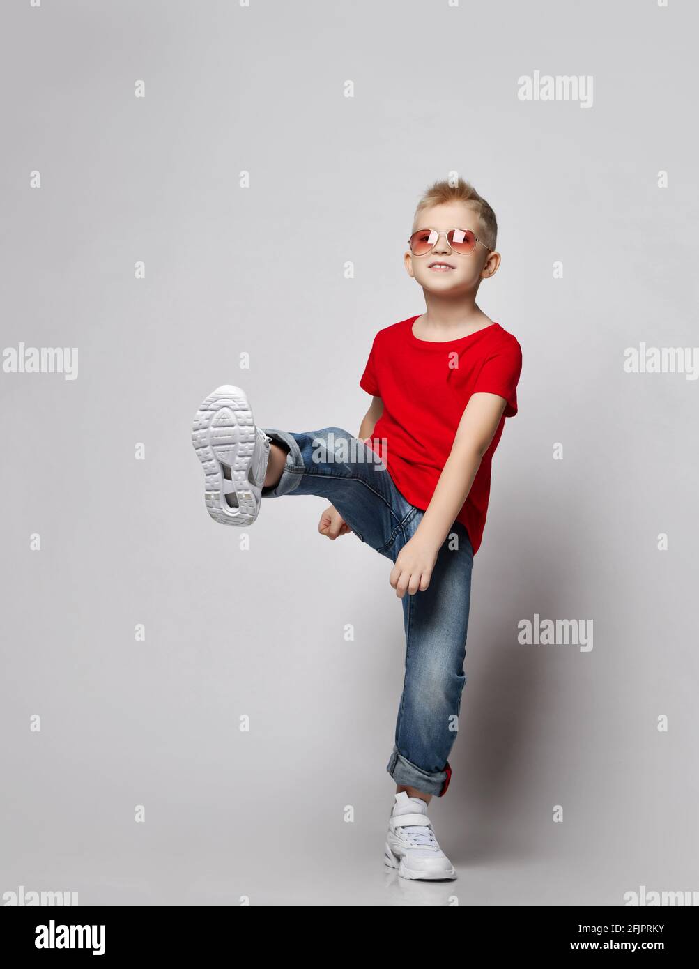 Frolic kid boy teenager in red t-shirt, blue jeans, white sneakers and sunglasses stands holding his foot up Stock Photo