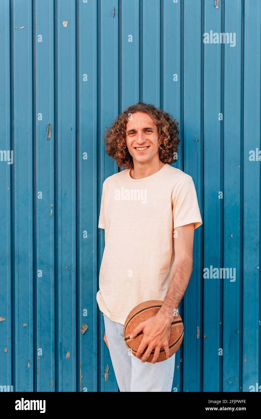 vertical portrait of a young caucasian man looking at camera smiling and holding a basketball. He has long red hair and wears casual summer clothes. It is leaning on a blue metal door Stock Photo