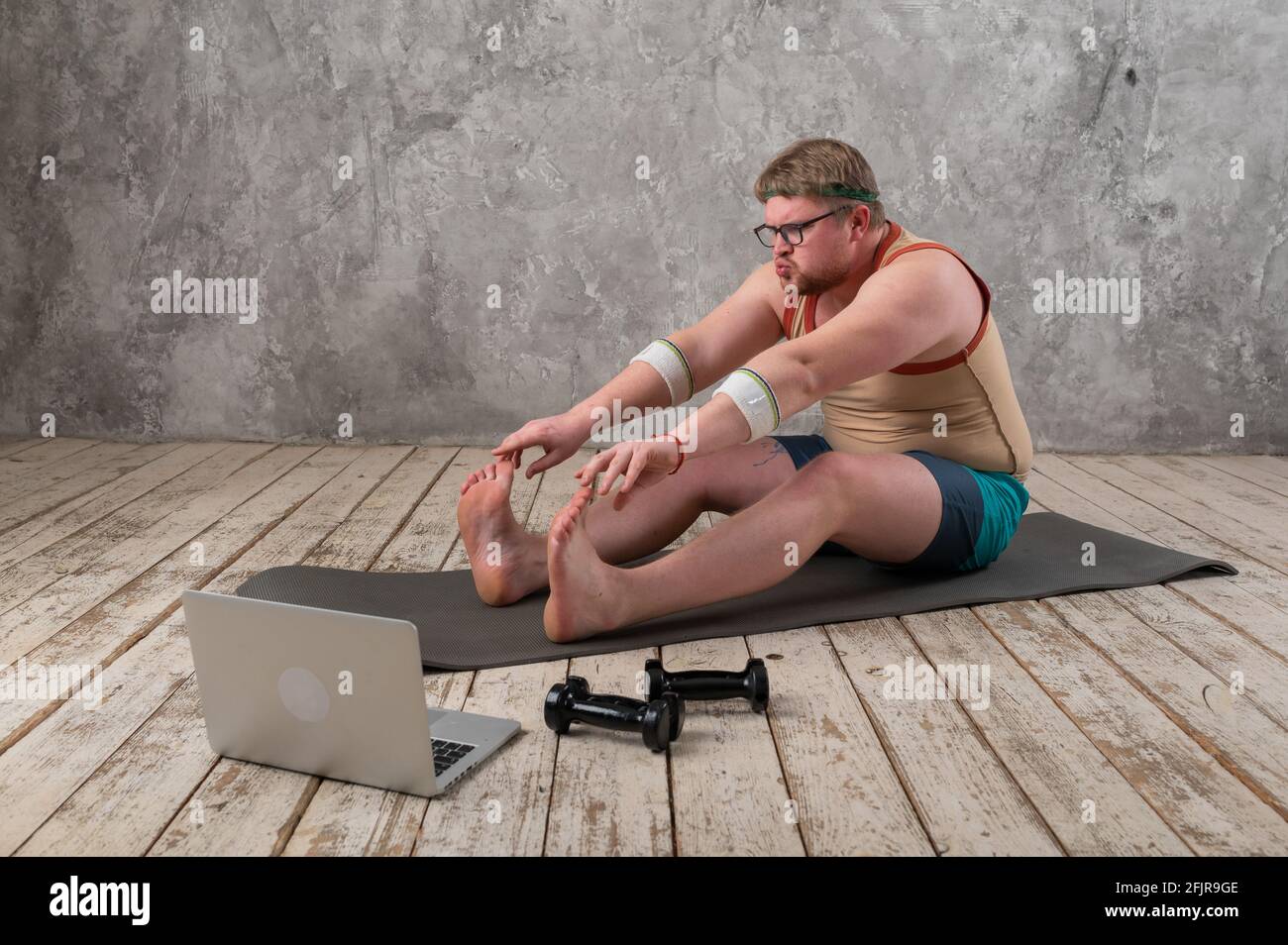 Funny fat man in sportswear doing sports exercises looking into a laptop. home workout  Stock Photo