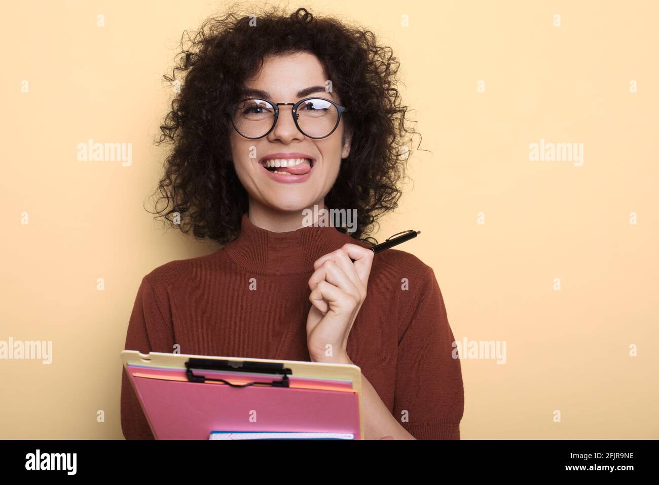 Adorable student with curly hair showing her tongue while holding folder on a yellow studio wall Stock Photo