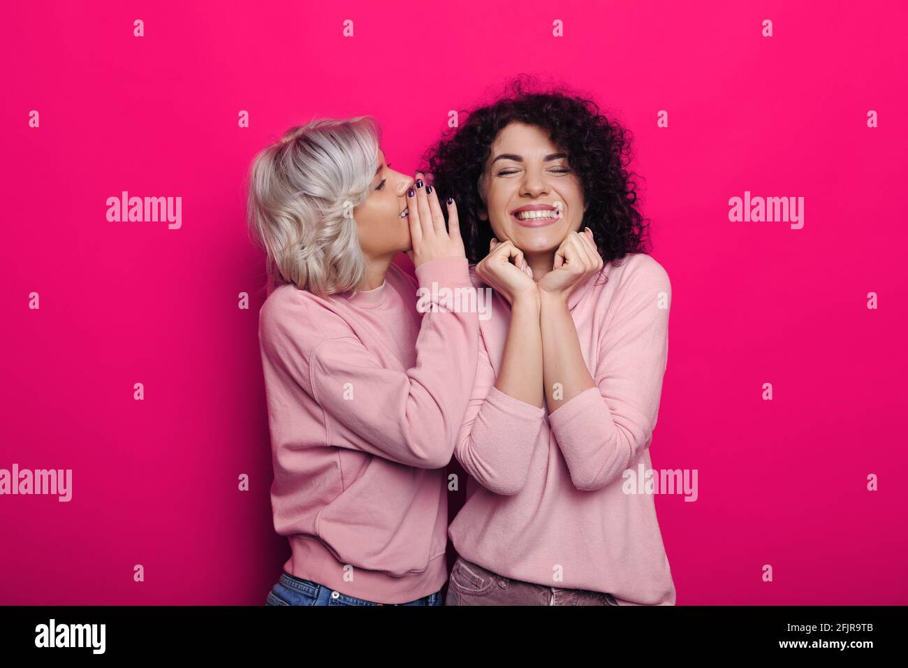 Blonde woman is whispering something to her brunette friend posing on a red studio wall Stock Photo