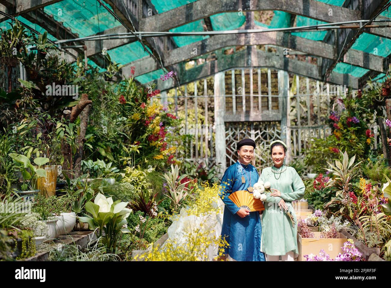 Happy young Vietnamese couple having wedding ceremony in greenhouse with many blooming flowers Stock Photo