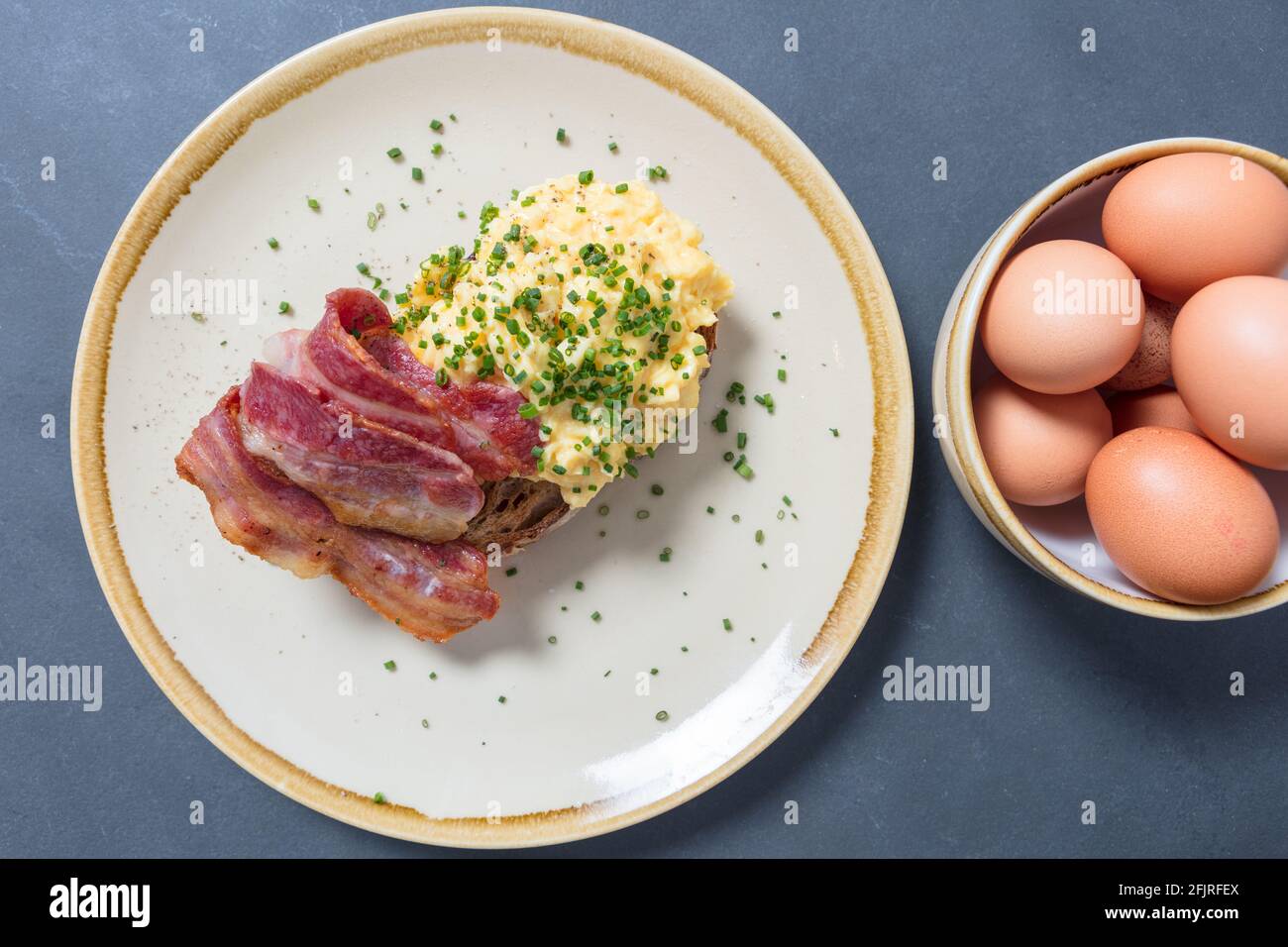 Scrambled eggs with bacon and chives on sourdough toast on plate with bowl of eggs to one side. Top down shot. Stock Photo