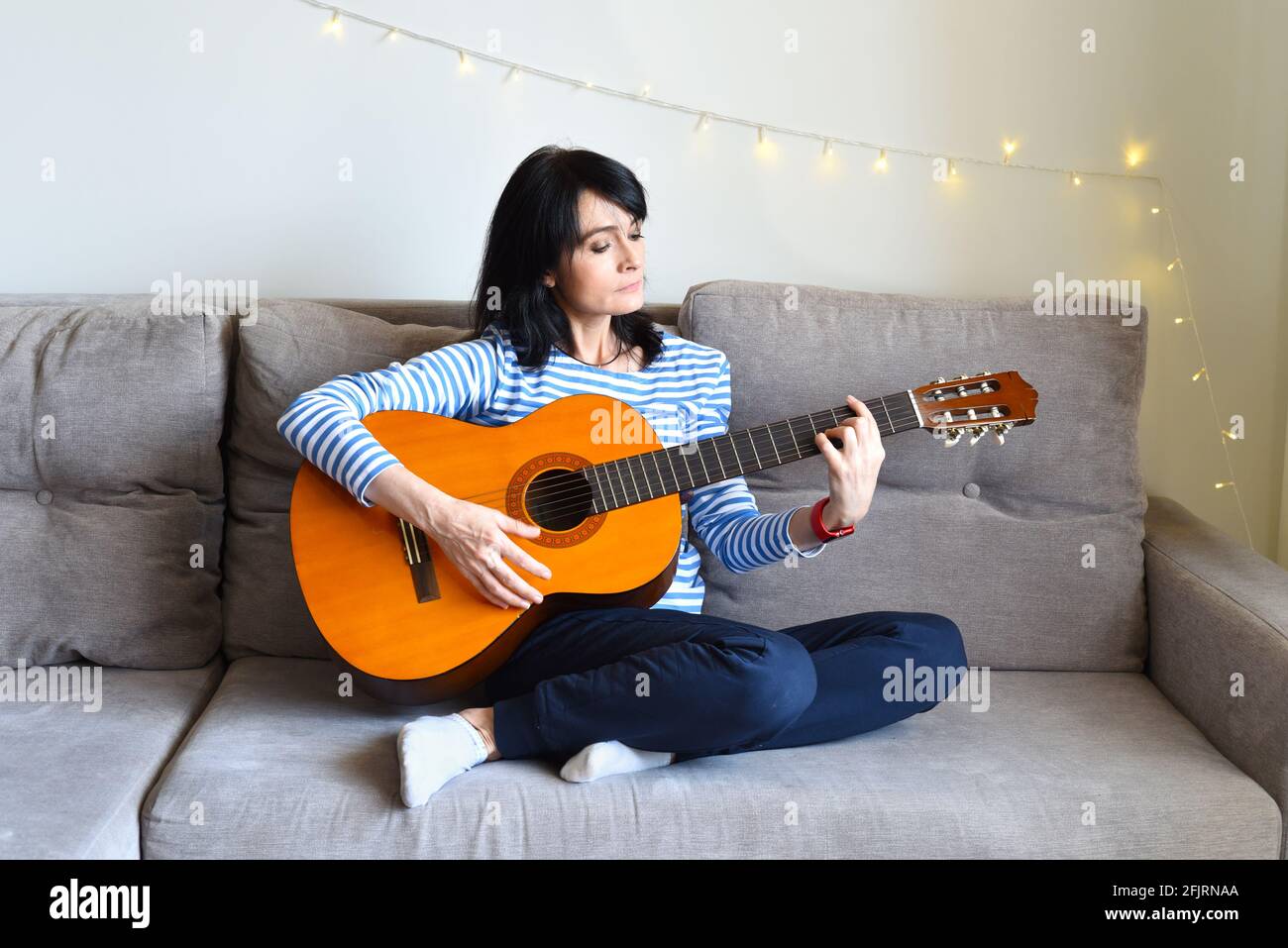 45 y.o. Russian woman playing guitar, sitting on a sofa at home. Stock Photo