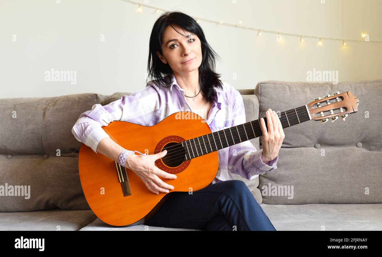 45 y.o. Russian woman playing guitar, sitting on a sofa at home. Stock Photo