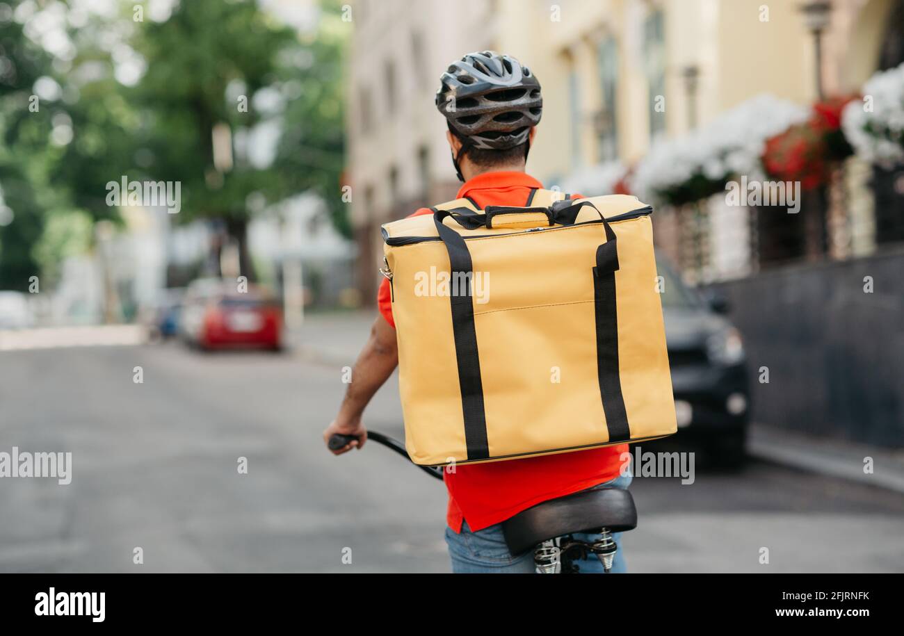 Young male courier in orange t-shirt, safety helmet with yellow thermo backpack, rides on bike on street road Stock Photo