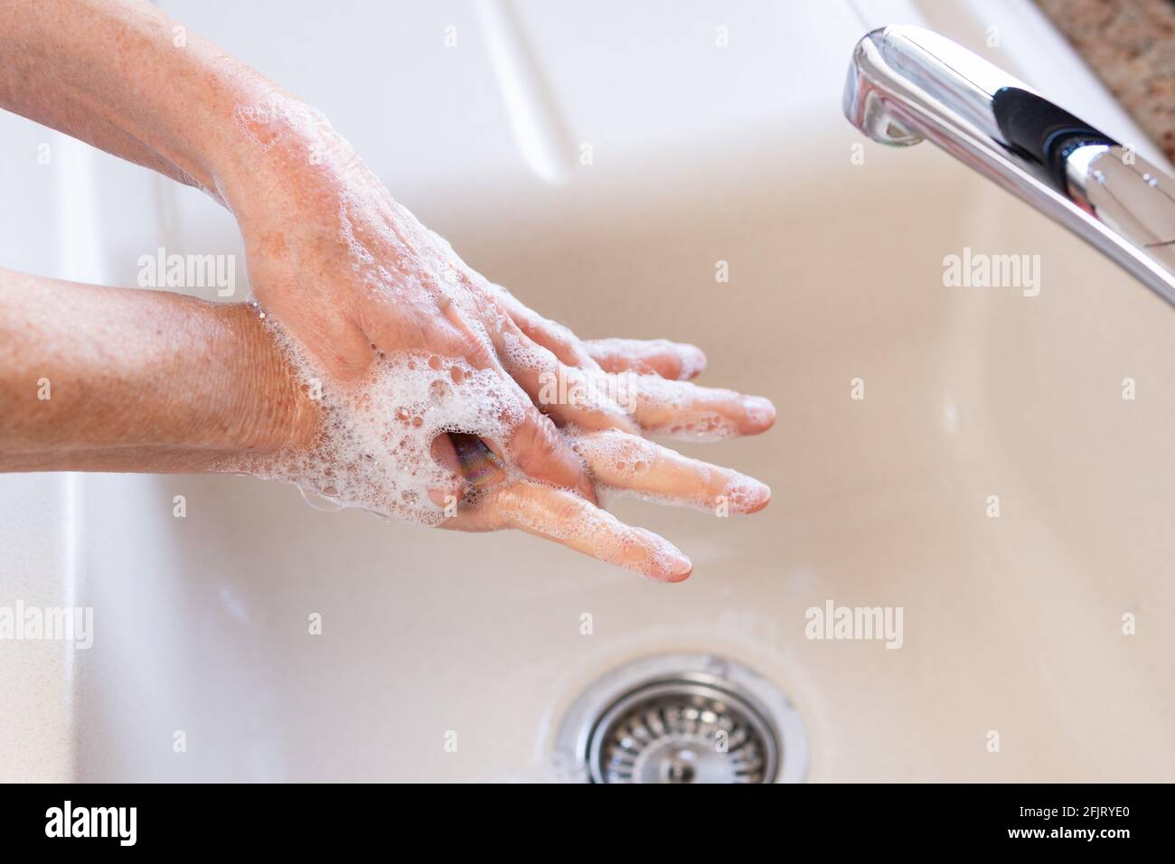 Elderly woman washing her hands with soap in kitchen sink. Fingers spread. Concept of personal hygiene for corona virus (covid-19) infection protection. Stock Photo