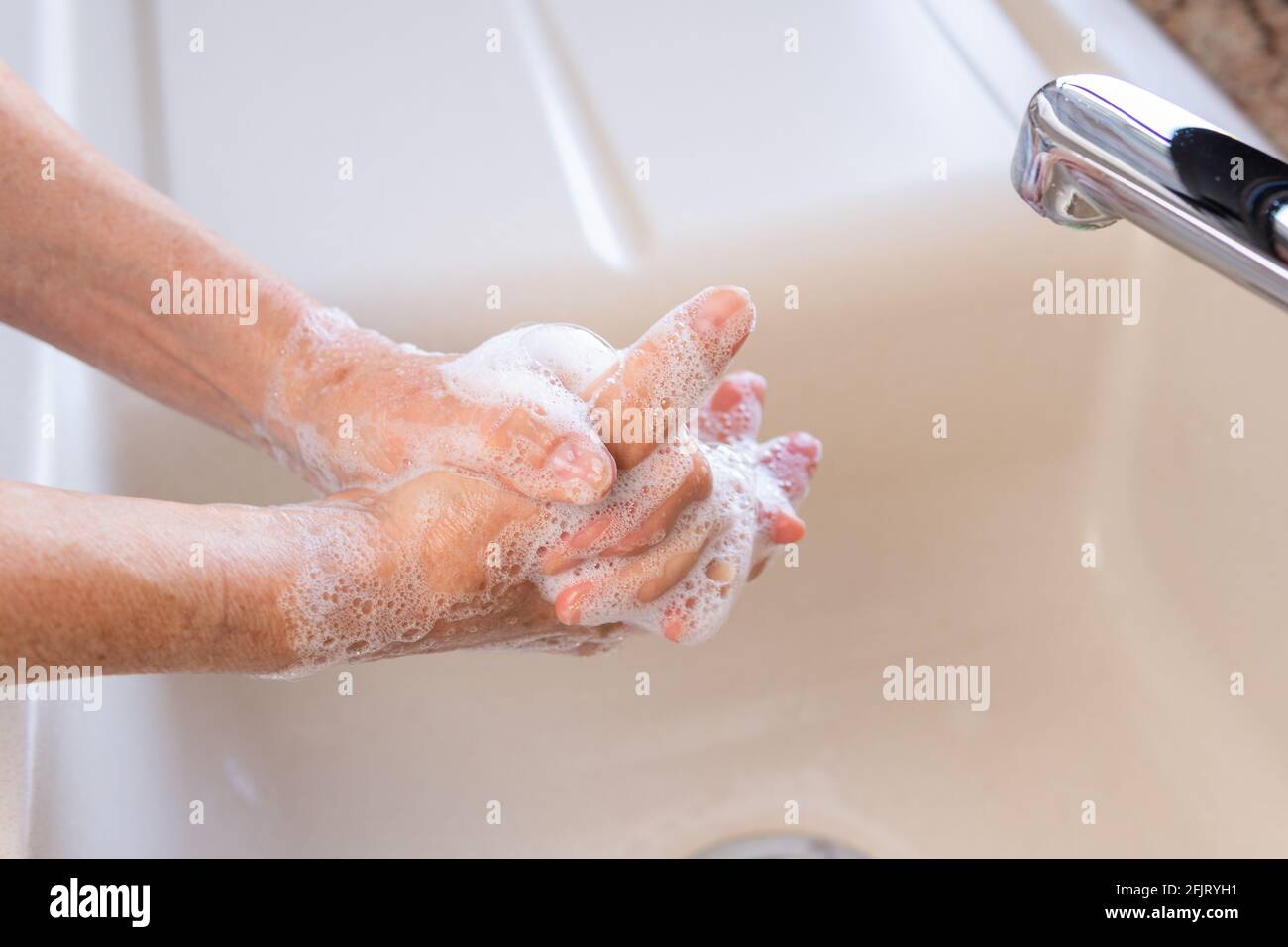 Elderly woman washing her hands with soap in kitchen sink under a faucet. Concept of personal hygiene for corona virus (covid-19) infection prevention. Stock Photo