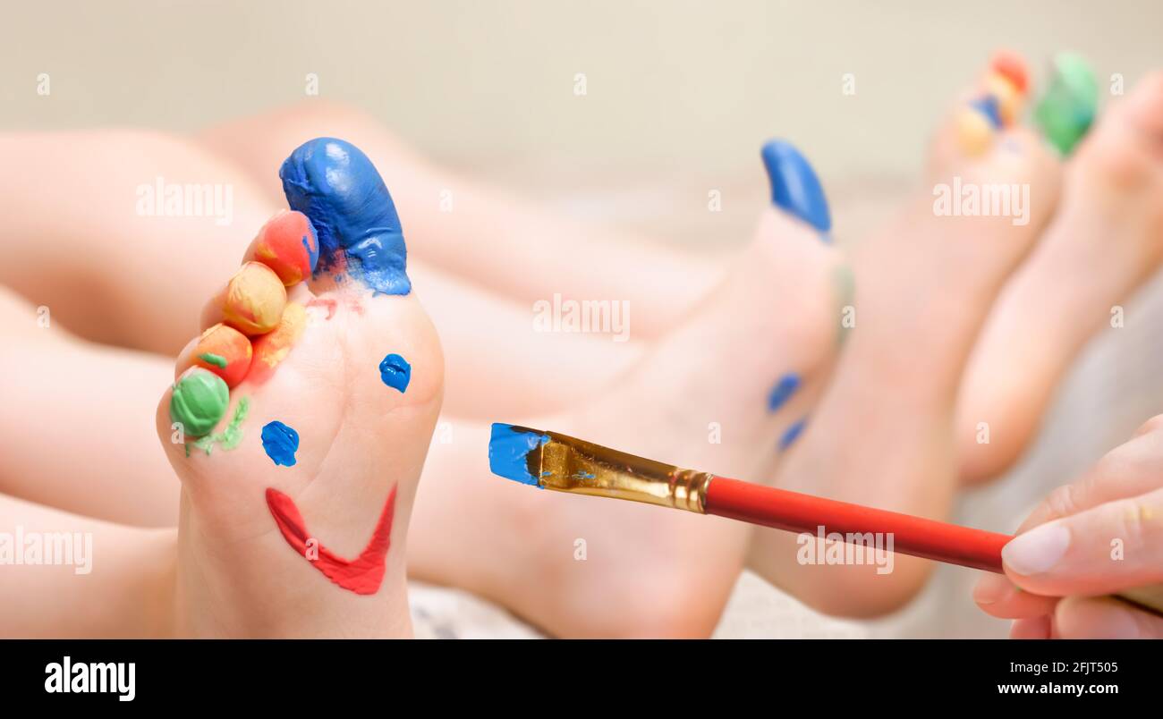 Close up hand brush drawing funny face on feet children with painted toes. Baby art therapy child painting face on legs kids paints smile on feet Stock Photo