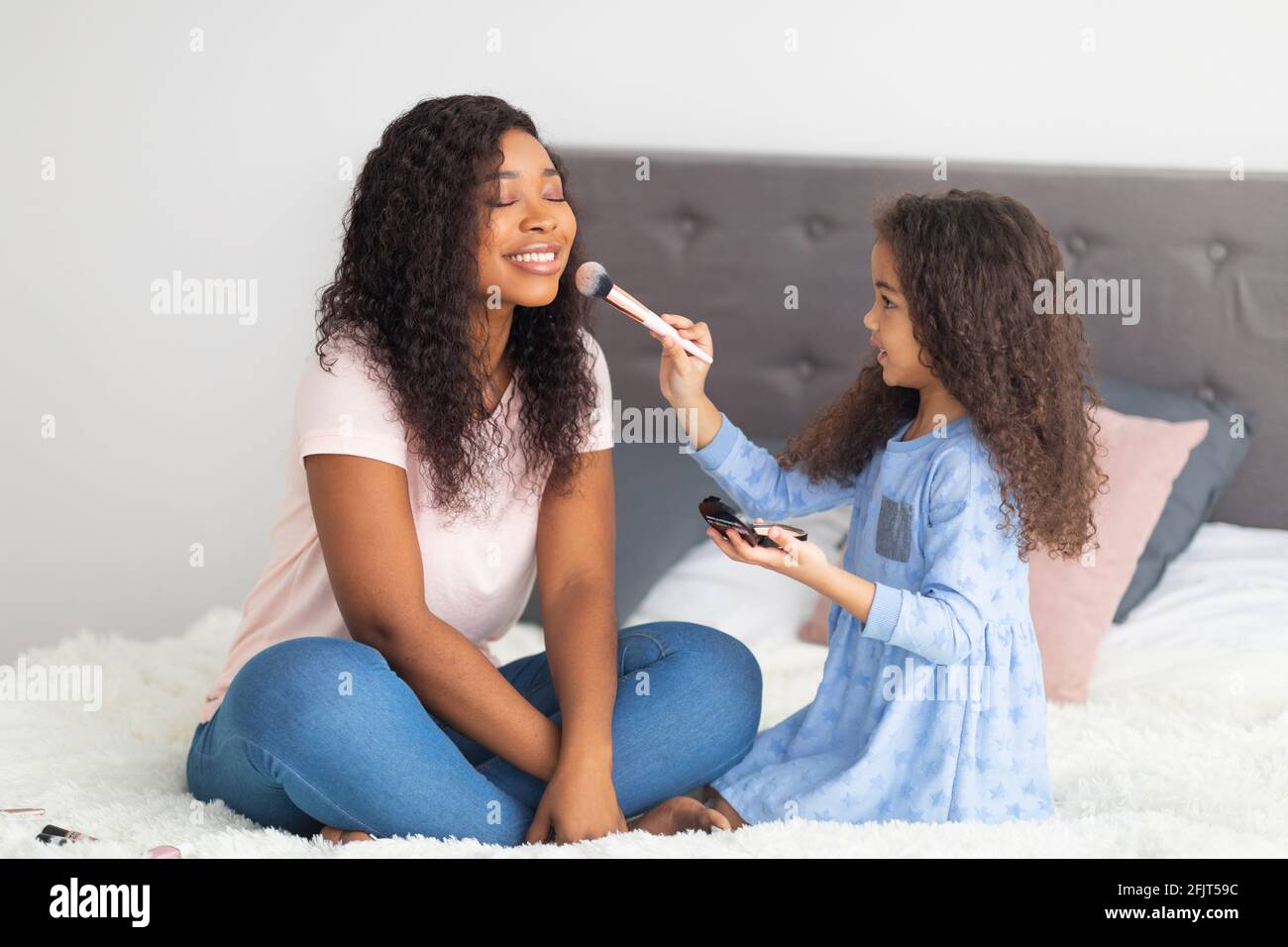 Joyful black girl applying makeup on her mom's face on bed at home