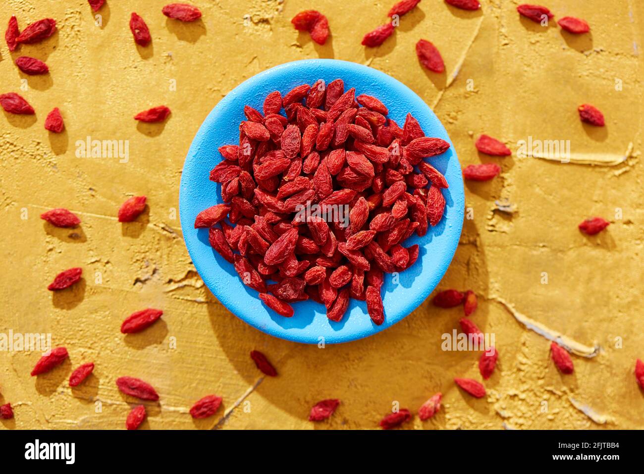 high angle view of some dried goji berries in a blue plate and sprinkled on a golden textured surface Stock Photo
