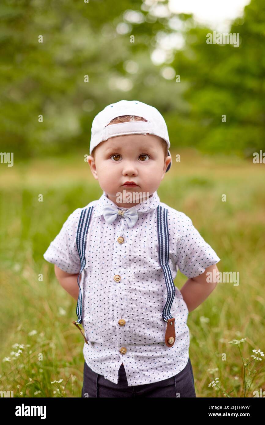 little serious boy in suspenders and a cap hides his hands behind back