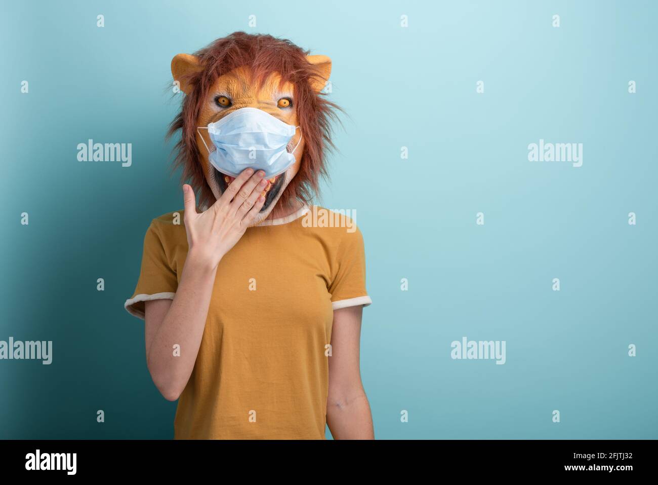 Young woman wearing lion and protection medical mask, covering mouth with hand, isolated on blue background. Stock Photo