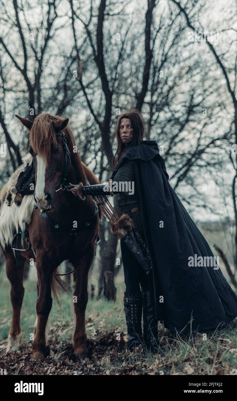 Woman in chain mail in image of medieval warrior holds her horse by the reins among forest. Stock Photo