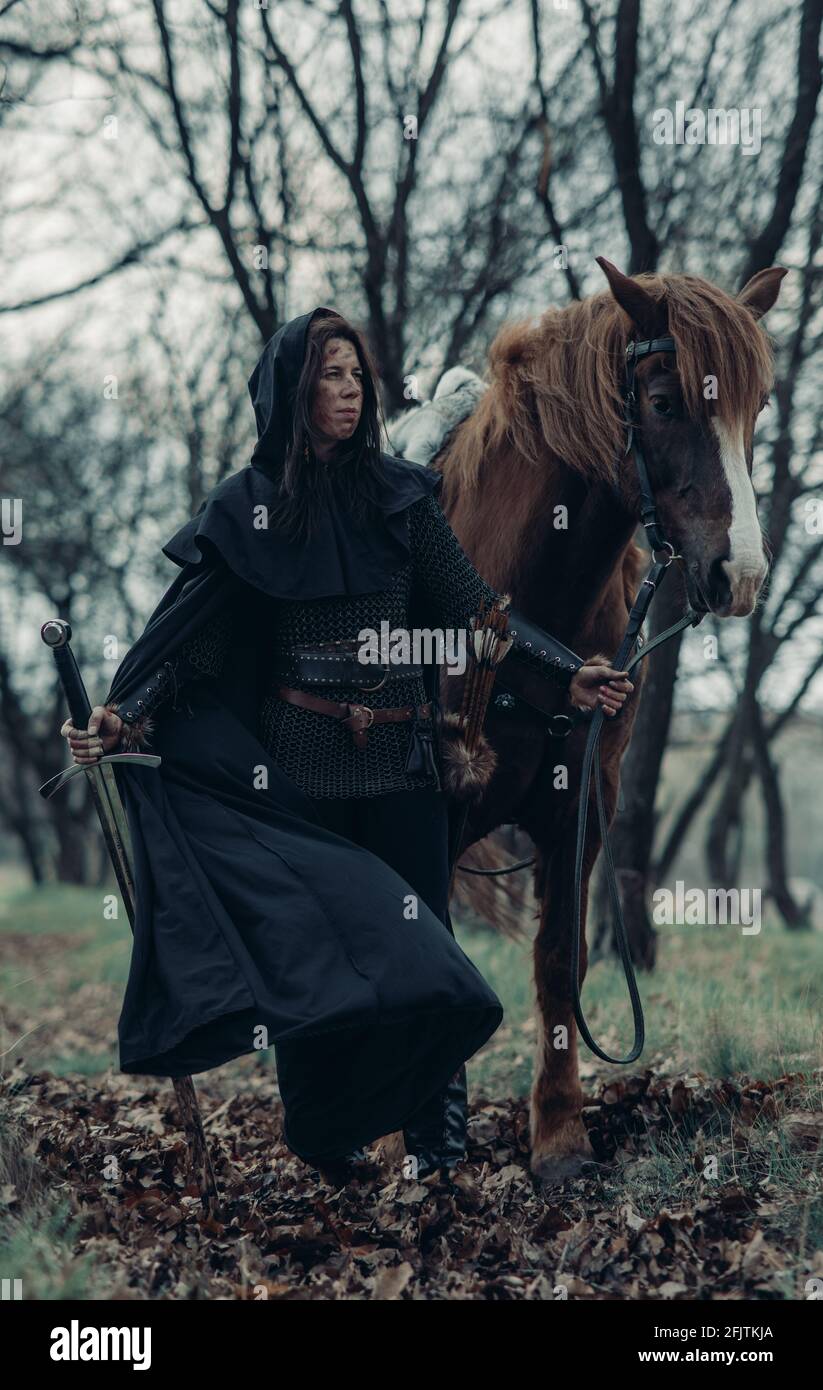 Woman in chain mail in image of medieval warrior leads her horse by the reins among forest with sword in her hand. Stock Photo