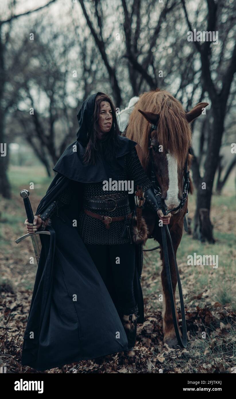 Woman in chain mail in image of medieval warrior leads her horse by the reins among forest with sword in her hand. Stock Photo