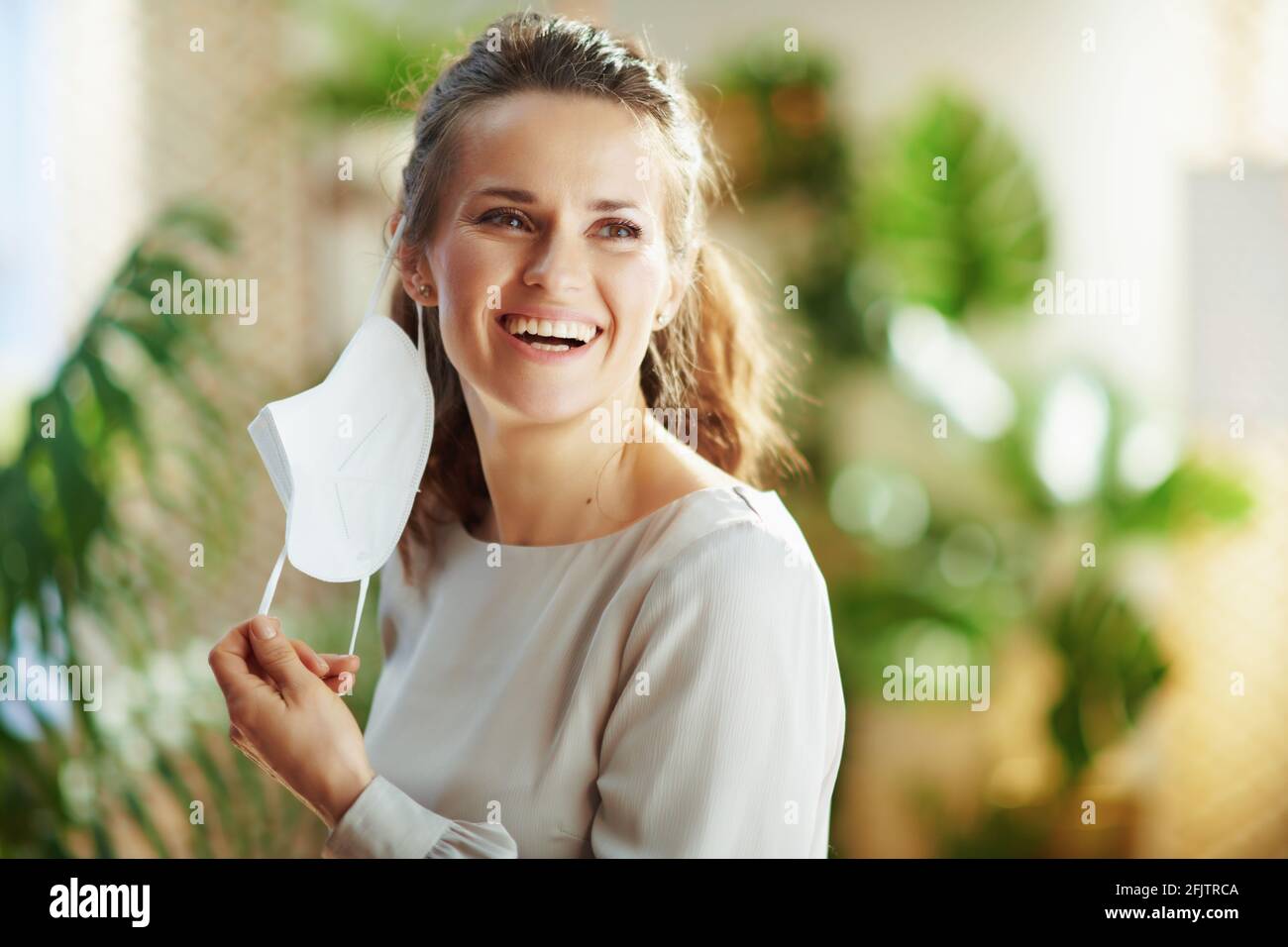 covid-19 pandemic. smiling modern woman in grey blouse taking off ffp2 mask in the modern house. Stock Photo