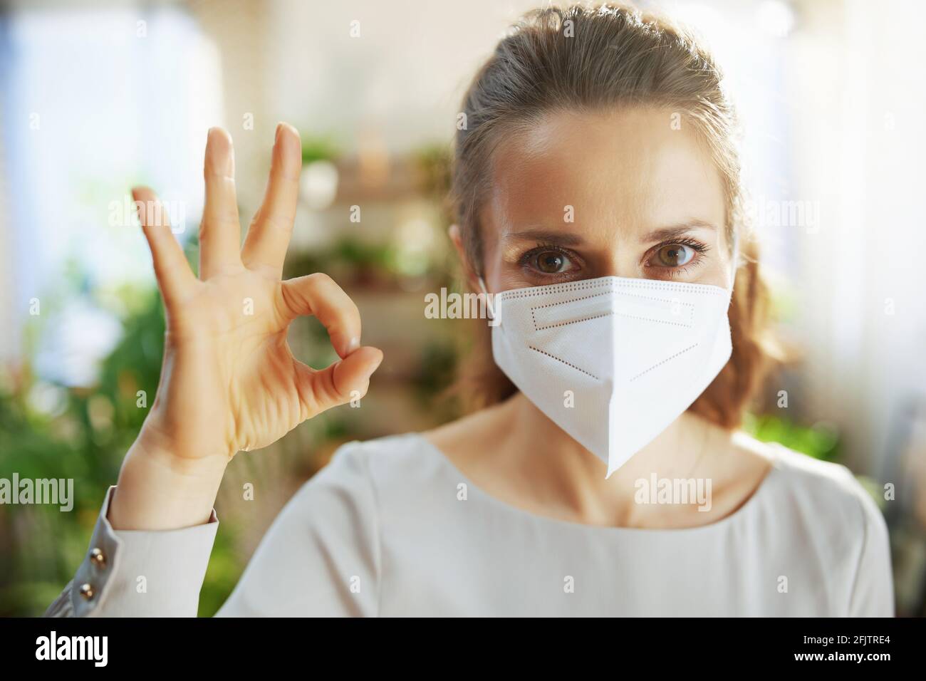 covid-19 pandemic. smiling trendy middle aged woman in grey blouse with ffp2 mask at modern home showing ok gesture. Stock Photo