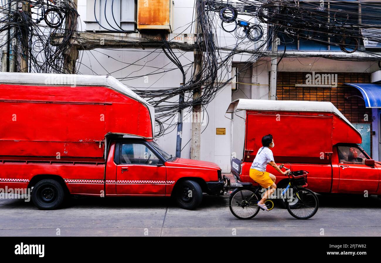 A woman cycles past two parked old red Mazda vans in the Chinatown area of Bangkok, Thailand Stock Photo