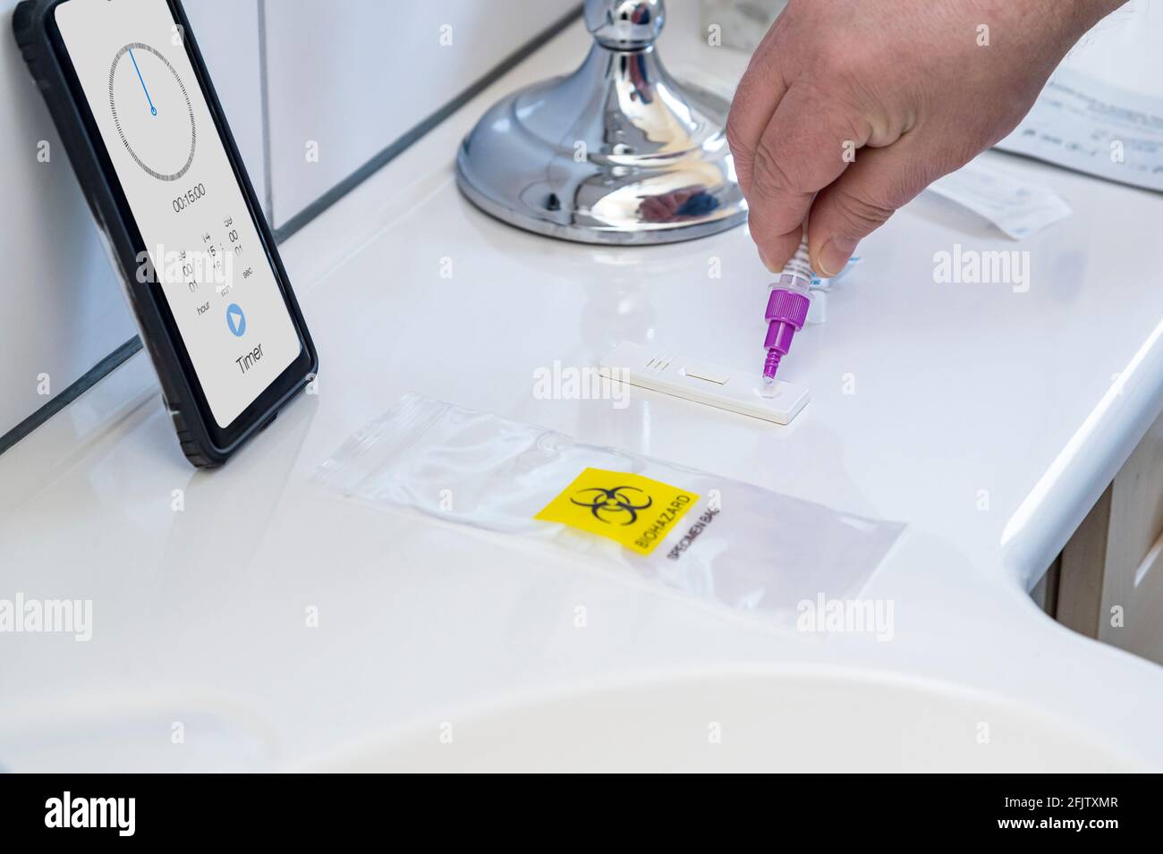 A man performs a Corona self-test. In the kit for SARS CoV-2 antigen test, he drips the test liquid onto the test strip. He is in a bathroom. A smartp Stock Photo