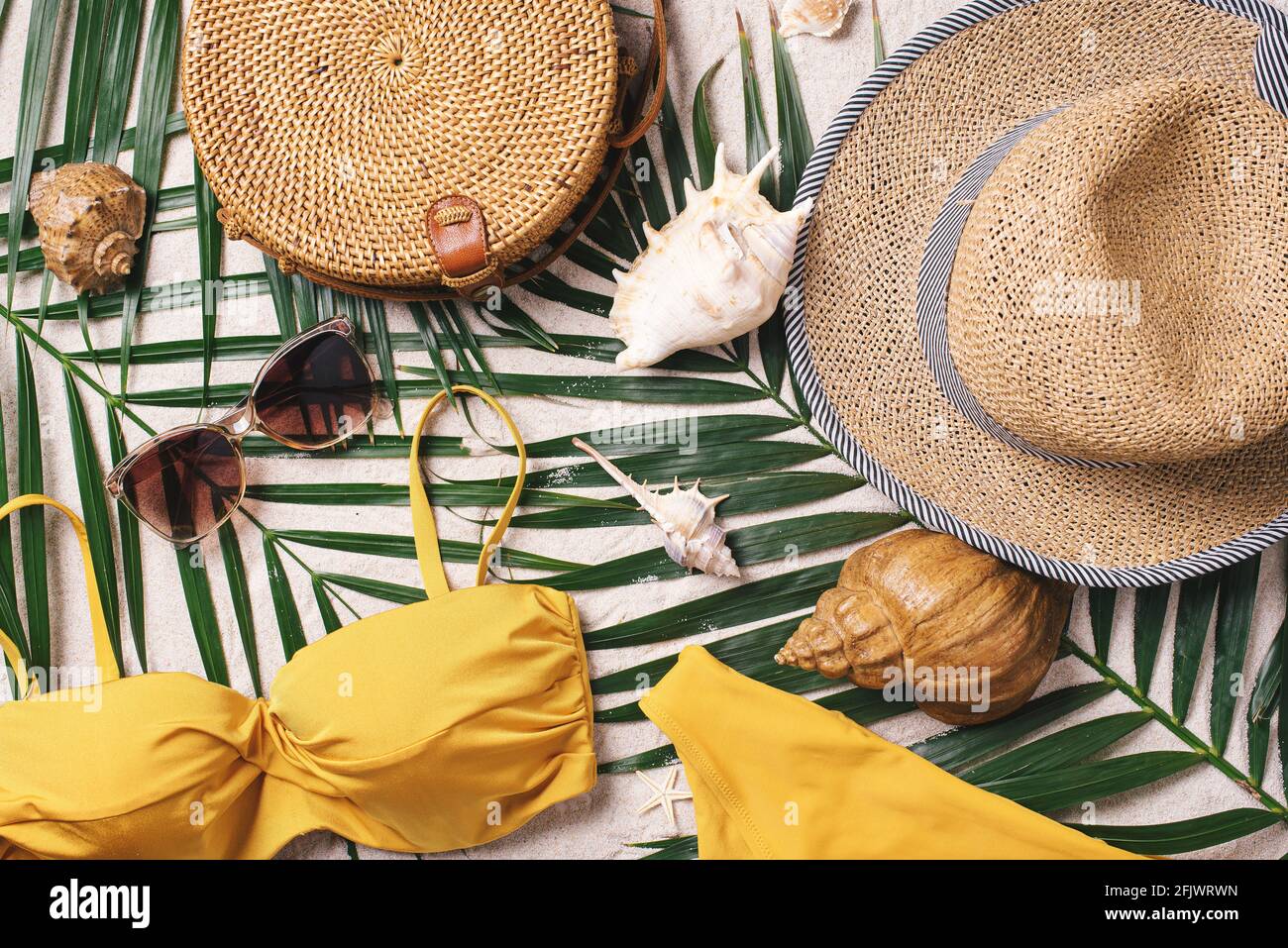 Female beach accessories: swimsuit, bikini, rattan bag, straw hat, shells, sunglasses, palm leaves on sand background. Exotic, tropical mood. Summer Stock Photo