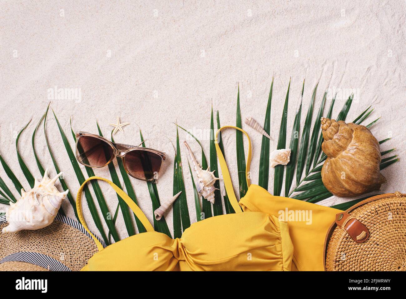 Woman's beach accessories: swimsuit, bikini, rattan bag, straw hat, shells, sunglasses, palm leaves on sand background. Exotic, tropical mood. Summer Stock Photo